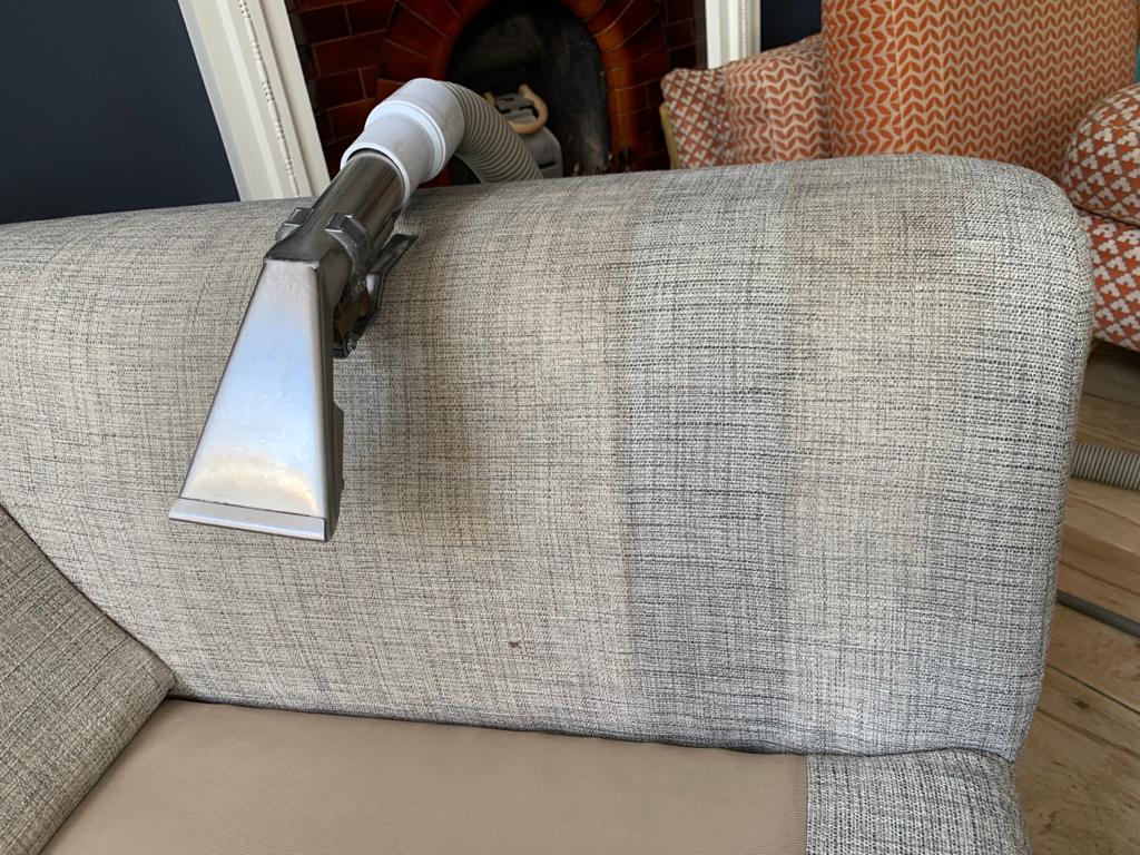 This is a photo of a sofa being cleaned by K&S Carpet Cleaning. It shows the head of the steam cleaning machine, and a before and after section of the sofa, showing one part that is dirty and one part that is clean.